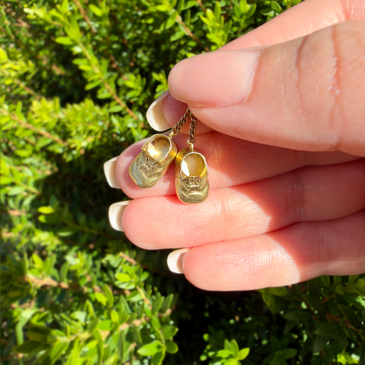 A close-up of a hand holding a pair of tiny gold miniature shoe charms against a background of green outdoor foliage.