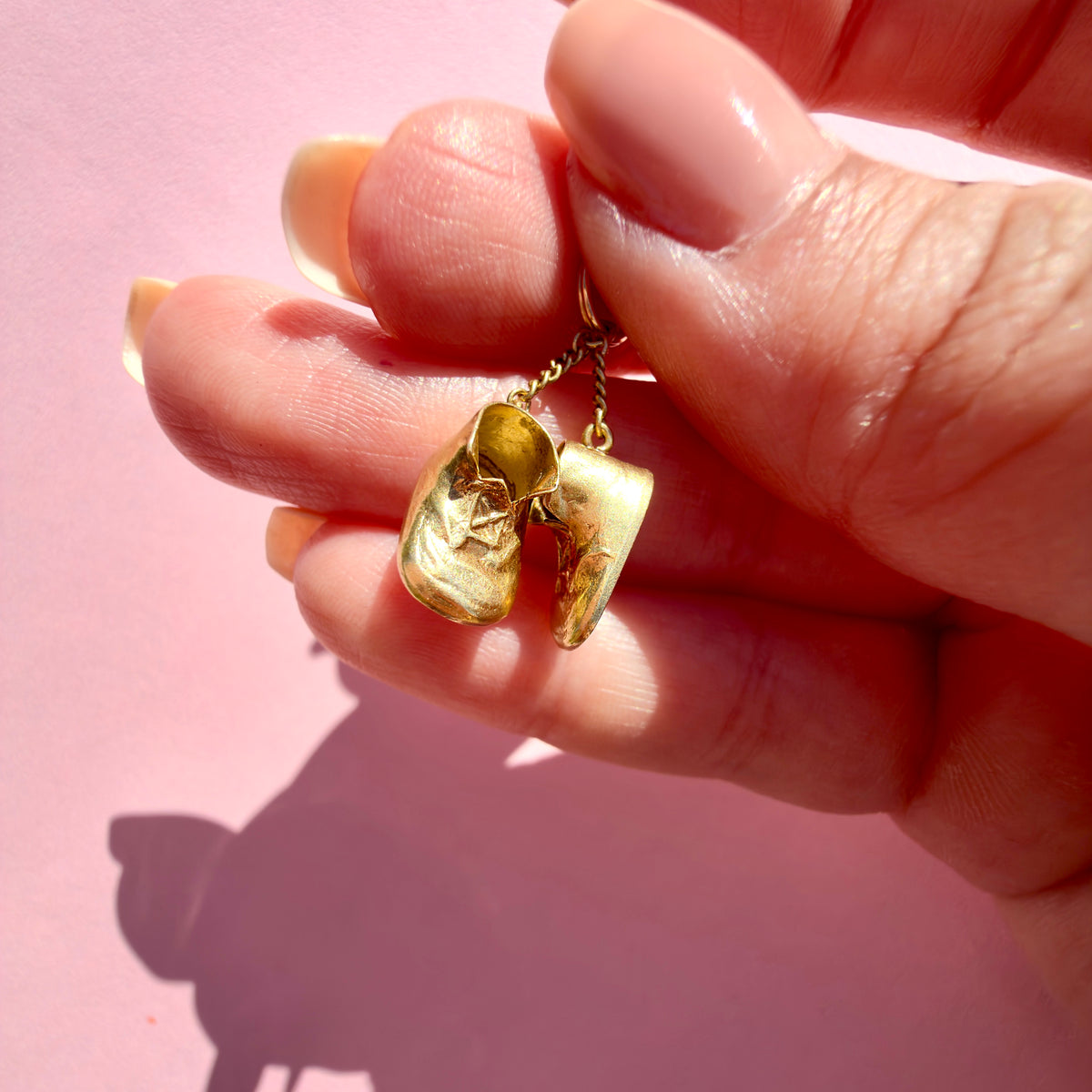 A close-up of a hand holding a gold pendant shaped like a pair of baby boots on a pink background.