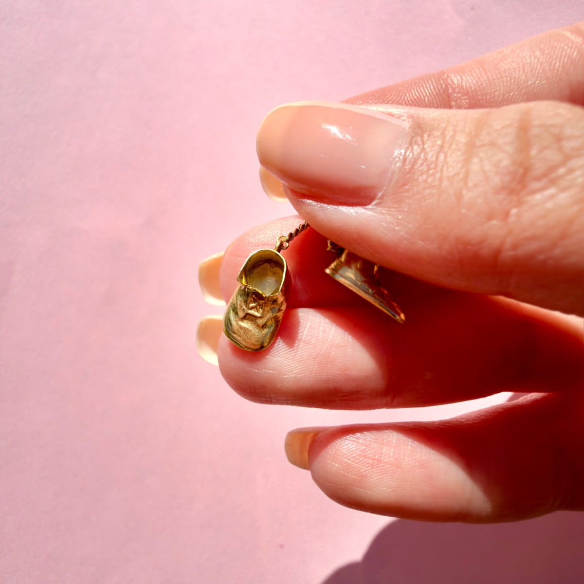 A close-up of a hand holding a tiny gold shoe charm against a solid pink background.