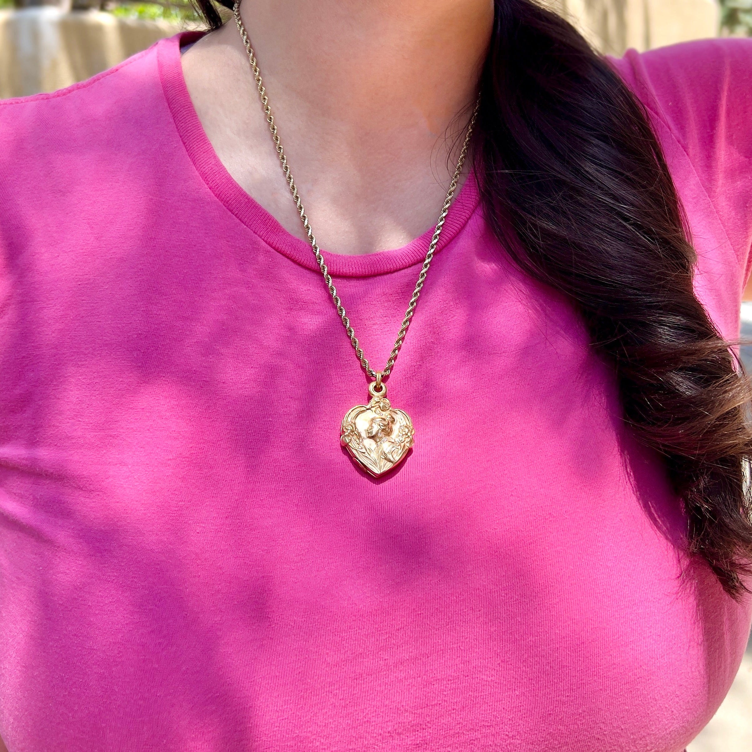 A close up of a person wearing a gold chain with an Art Nouveau heart-shaped locket with gold detailing, featuring a central figure with a bun and flowers on the front.