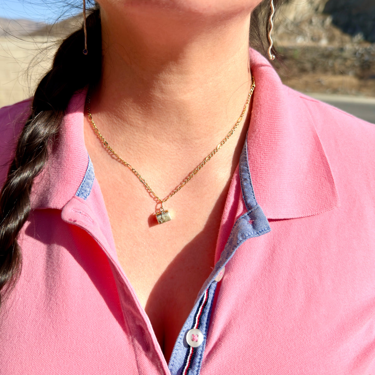 A close-up of a woman wearing a delicate gold chain necklace with a small gold mailbox charm, paired with a pink polo shirt.