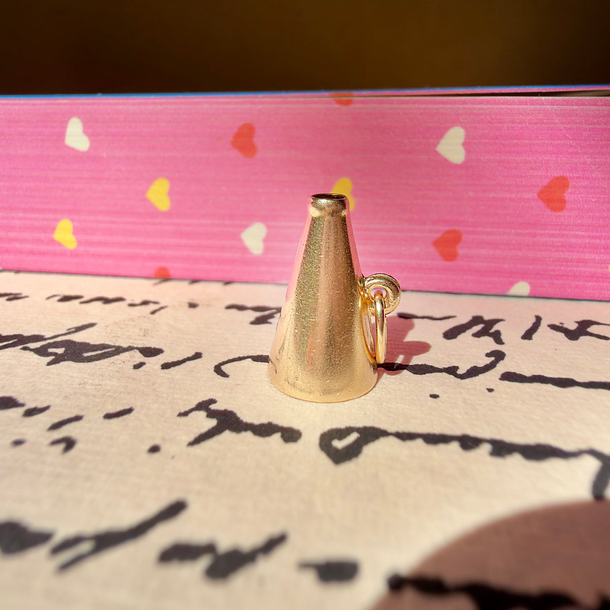 A megaphone charm with a small attached ring, standing upright on cream-colored paper with cursive writing, with a pink heart-patterned book in the background.