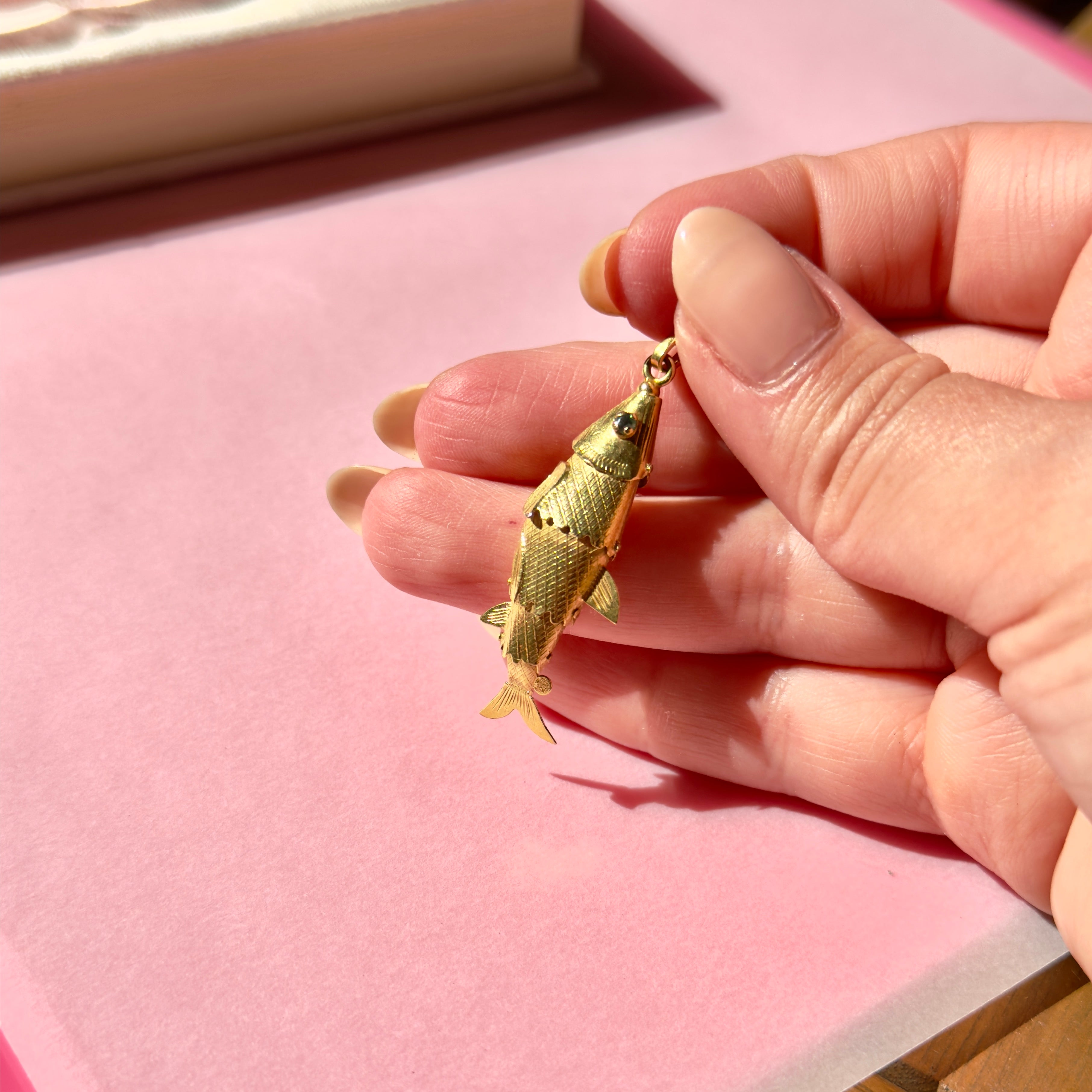 Gold fish-shaped pendant held between fingers on a pink surface