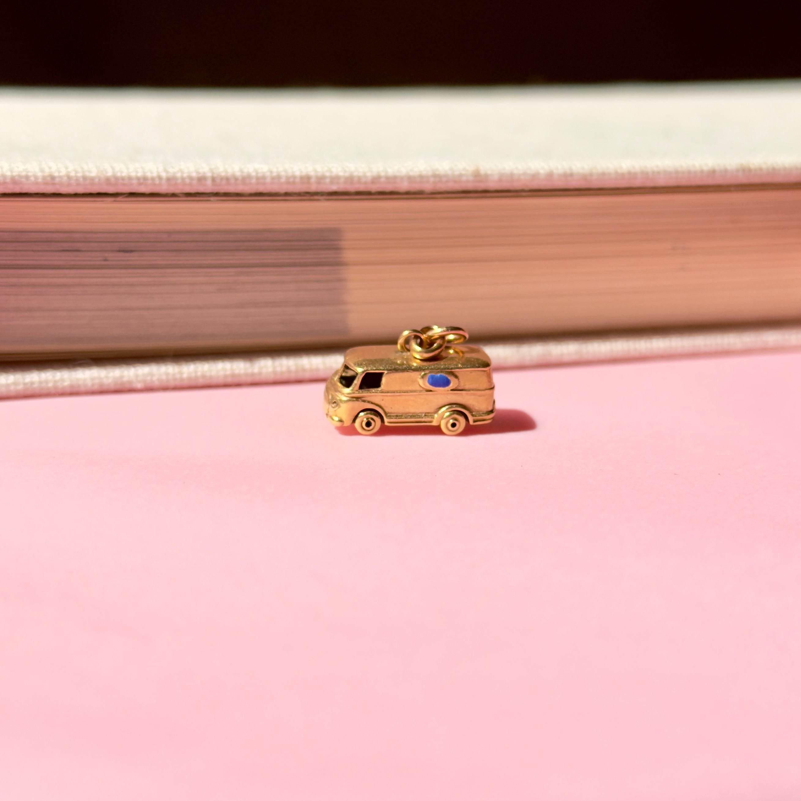 Vintage gold microbus  charm with a light blue enamel window on a pink background in front of a book