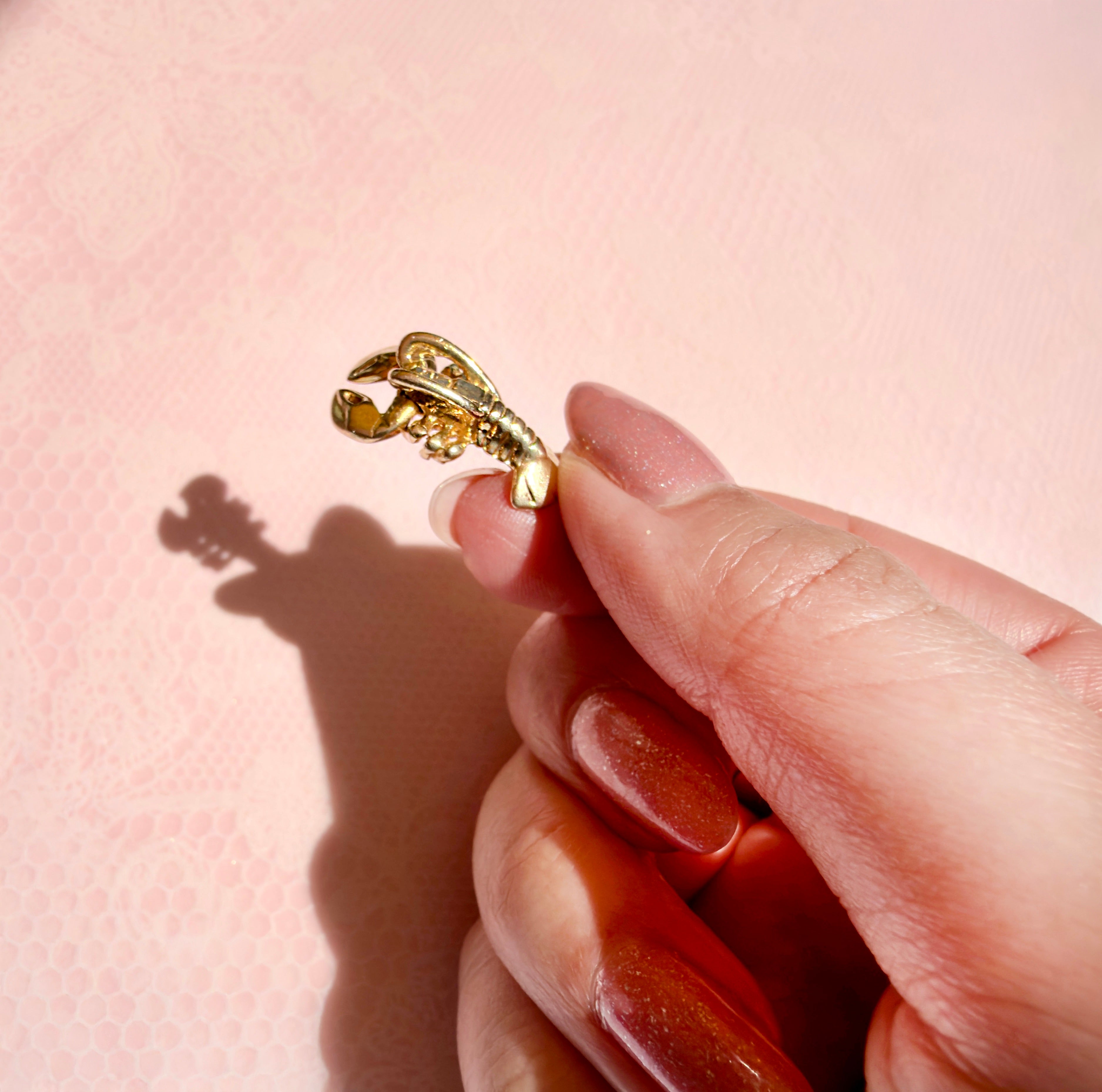 A hand holding a gold lobster charm on a pink background