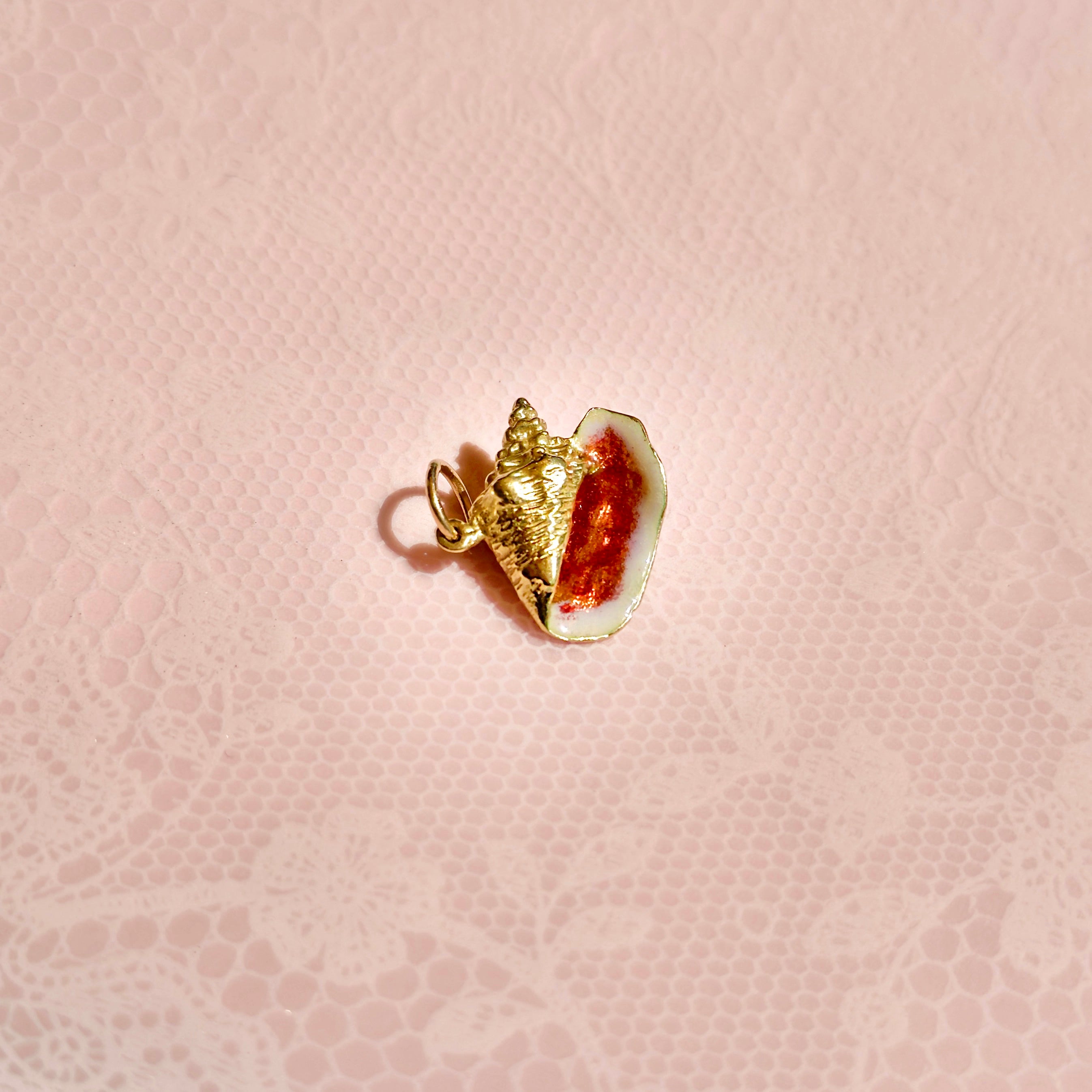 An 18K yellow gold vintage conch shell charm with orange enamel against a pink background.
