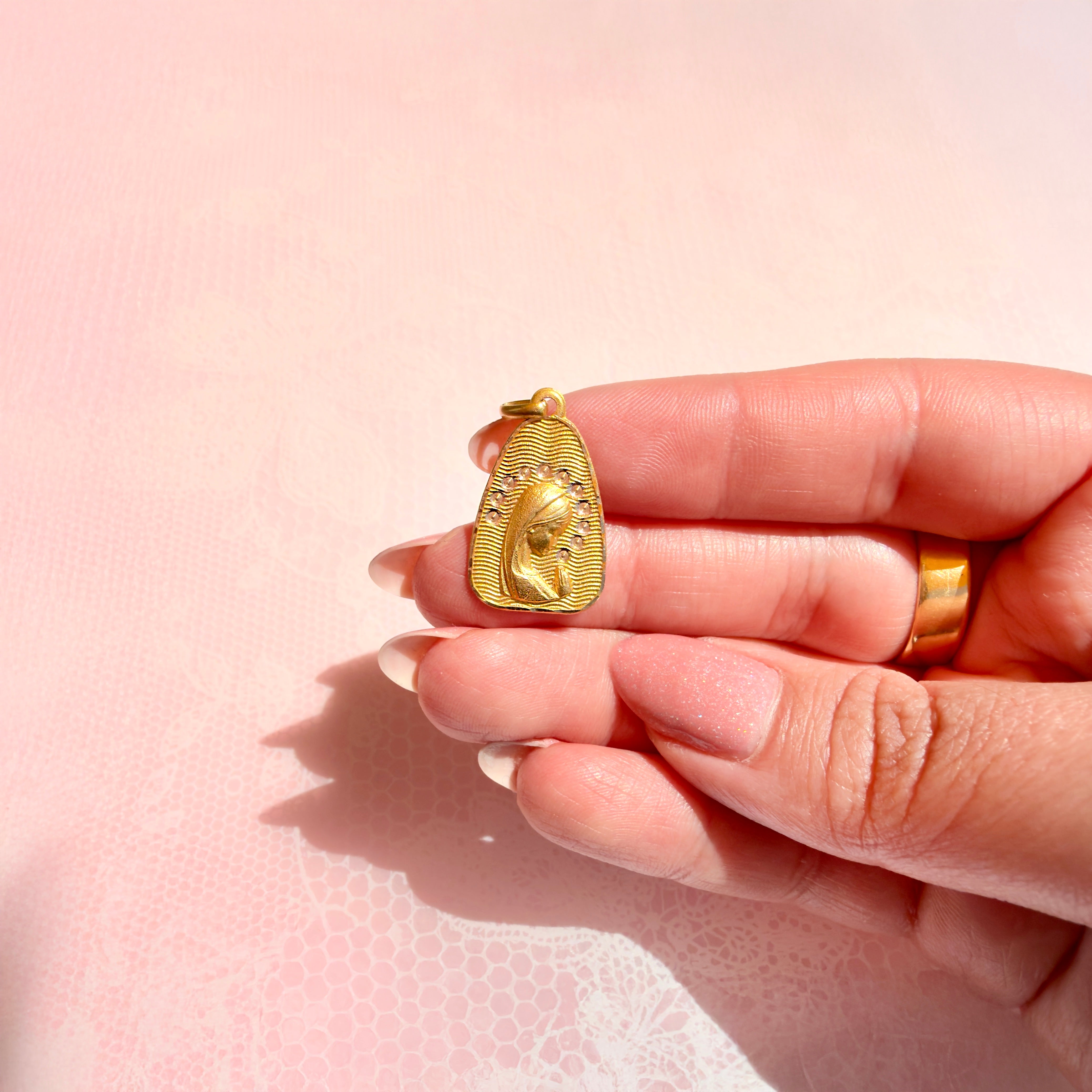 A hand holding a gold pendant with a religious design against a pink background.
