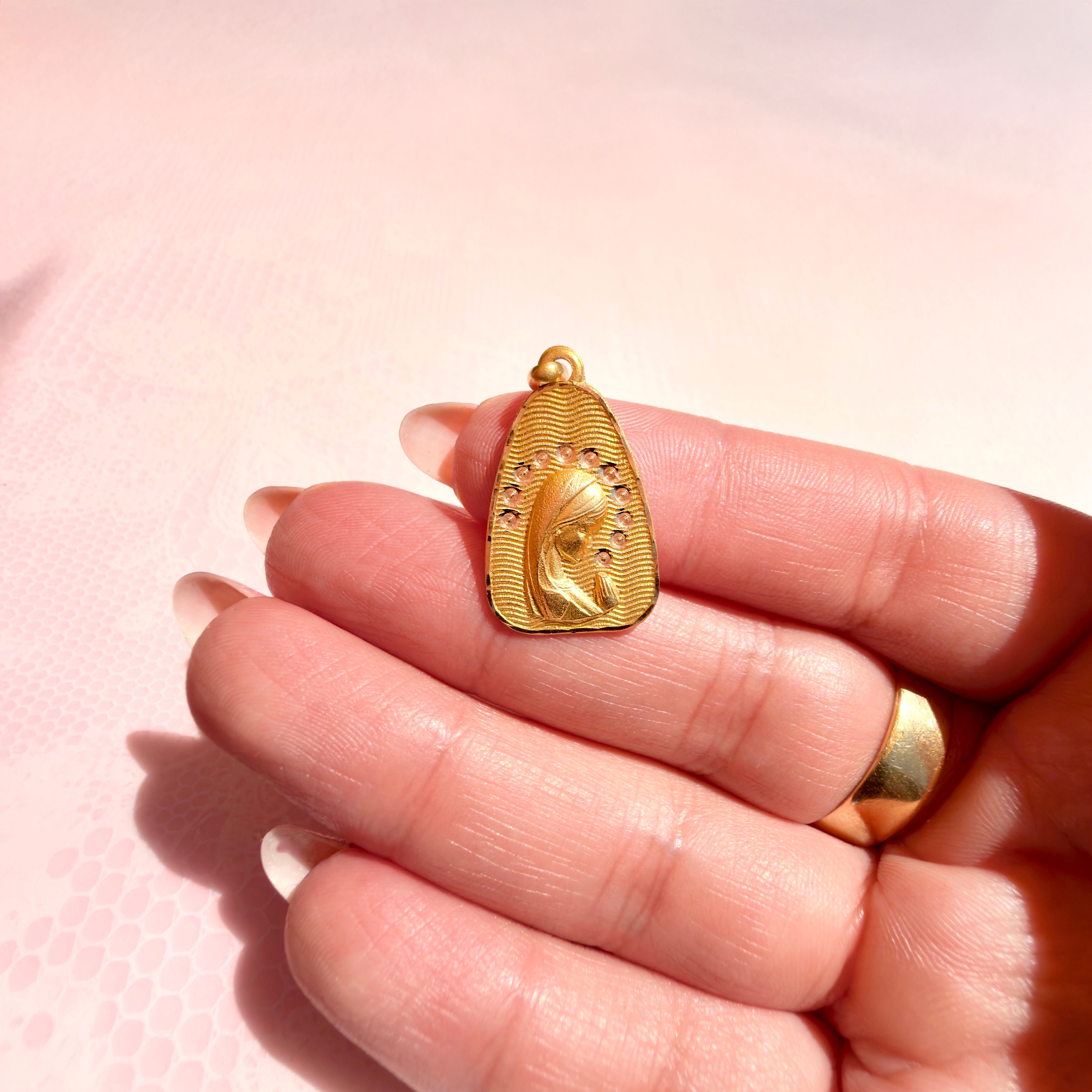 A hand holding a gold pendant with a religious design against a pink background.
