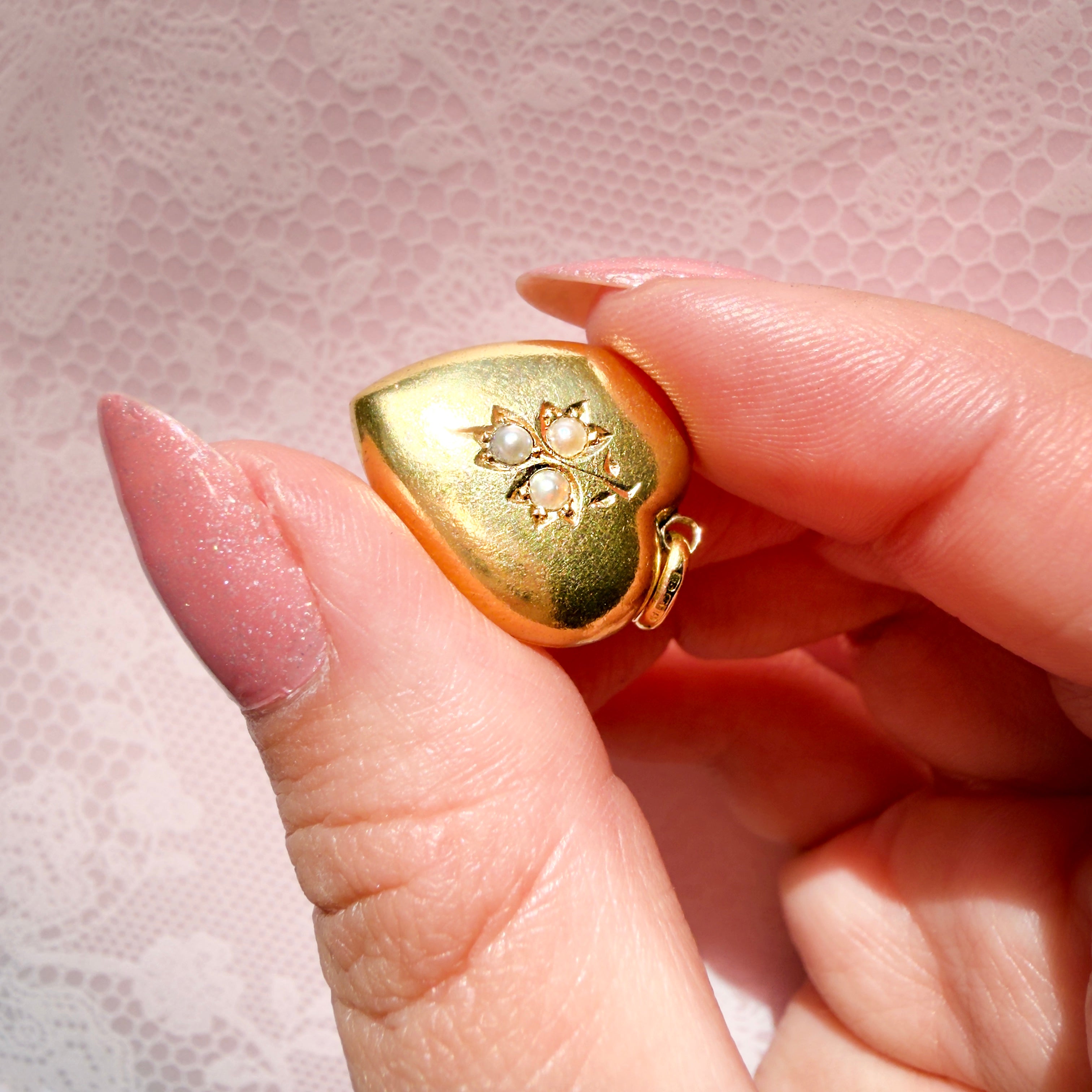 Hand holding a gold heart-shaped pendant with pearls on a pink background