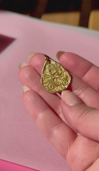 Gold pendant with a car design held in a hand against a pink background
