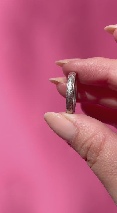 Two fingers holding a vintage platinum ring with intricate engraving against a pink background