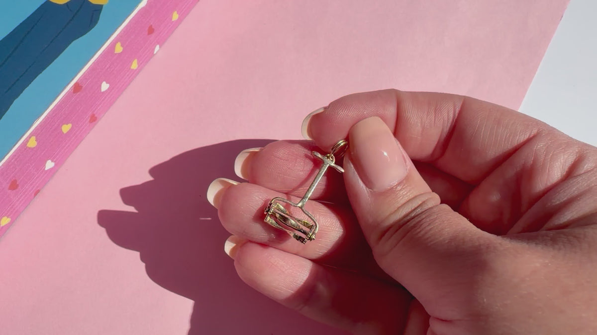 A close-up of a hand holding a small gold lawn mower charm against a pink background.