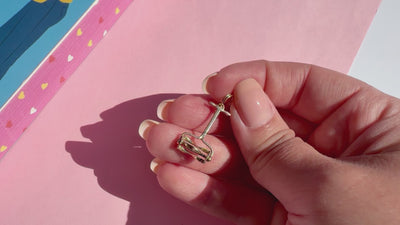 A close-up of a hand holding a small gold lawn mower charm against a pink background.