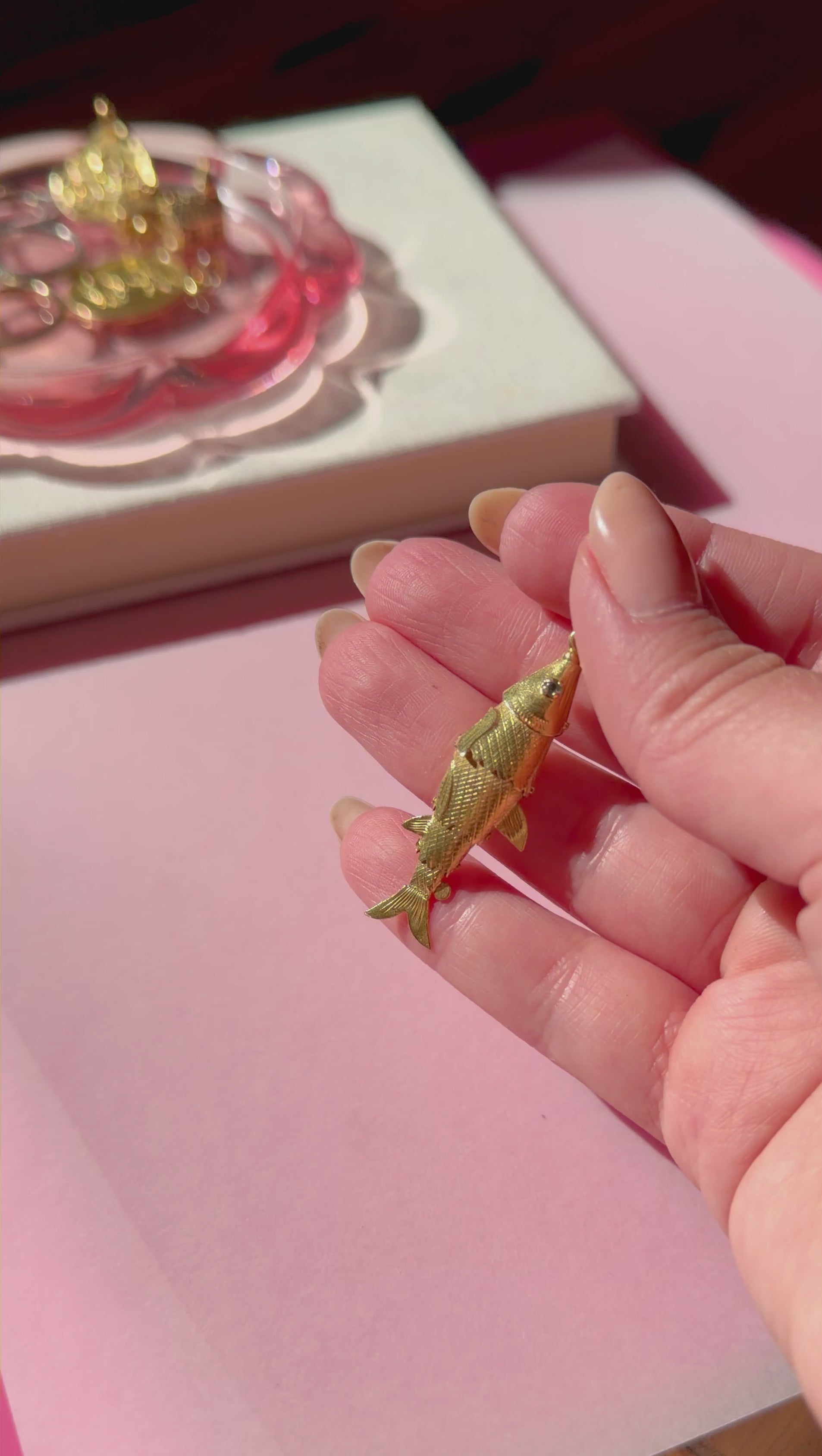 Gold fish-shaped pendant held between fingers on a pink surface