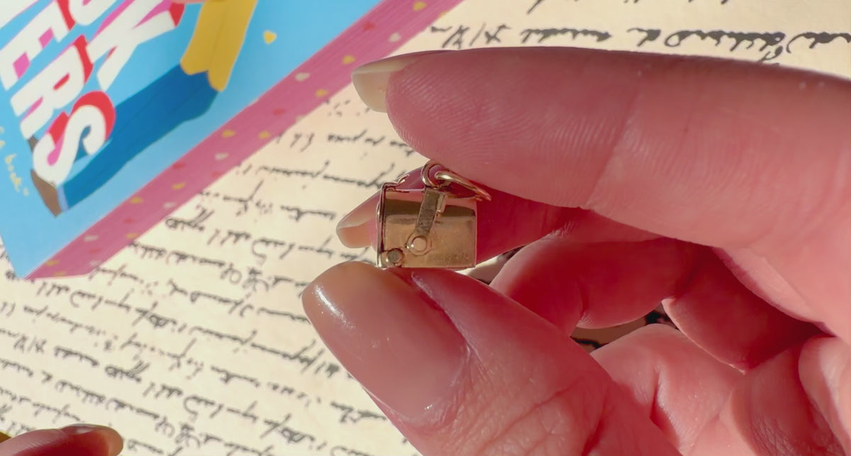A close-up of a hand holding a small gold mailbox charm against a blurred colorful background.