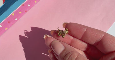 A small gold charm shaped like a stroller resting on a person's fingers, with a blurred pink background.